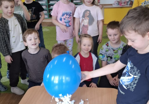 Dzieci stoją przy stoliku i obserwują eksperyment z balonem i kawałkami papieru.