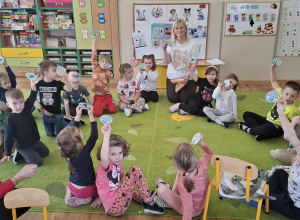 Dzieci siedzą na dywanie i pokazują odznaki z okazji Dnia Dinozaura.