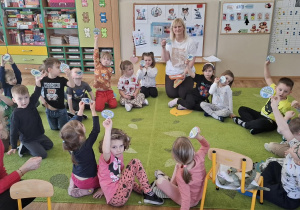 Dzieci siedzą na dywanie i pokazują odznaki z okazji Dnia Dinozaura.