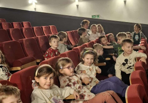 Dzieci znajdują się w sali kinowej i siedzą na fotelach.