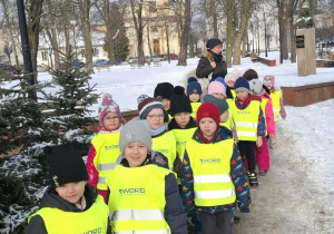 Dzieci stoją parami w odblaskowych kamizelkach w parku miejskim.