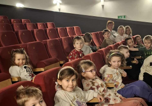 Dzieci znajdują się w sali kinowej i siedzą na fotelach.