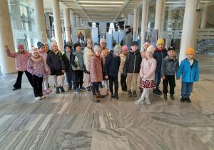 Grupa dzieci ubrana w kurtki i czapki stoi przed wejściem do teatru