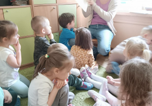 Dzieci siedzą na dywanie i oglądają obrazki w książeczce, którą czytała mama