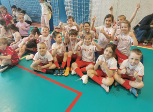 Uśmiechnięta reprezentacja przedszkola siedząc na boisku prezentuje kciuki do góry.