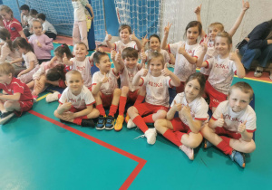 Uśmiechnięta reprezentacja przedszkola siedząc na boisku prezentuje kciuki do góry.