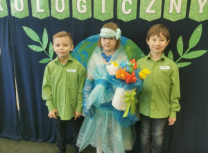 Zespół, w składzie: dziewczynka i dwaj chłopcy w zielonych koszulach, stoi na tle napisu „Turniej ekologiczny”, a dziewczynka ubrana jest w sukienkę wykonaną z folii z recyklingu i w dłoni t