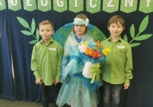 Zespół, w składzie: dziewczynka i dwaj chłopcy w zielonych koszulach, stoi na tle napisu „Turniej ekologiczny”, a dziewczynka ubrana jest w sukienkę wykonaną z folii z recyklingu i w dłoni t