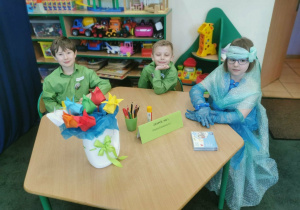 Dwaj chłopcy i dziewczynka siedzą przy stoliku konkursowym – są reprezentacją Miejskiego Przedszkola nr 1 na Turnieju Ekologicznym.