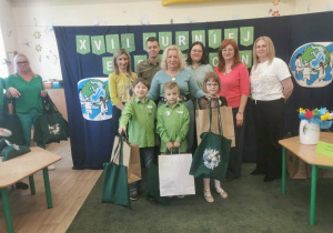 Trzyosobowy zespół dzieci reprezentujący przedszkole stoi wraz z organizatorami konkursu, jury, przedstawicielem nadleśnictwa oraz nauczycielką grupy, a dzieci prezentują nagrody konkursowe.