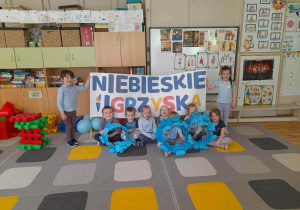 Grupa dzieci siedzi na dywanie i prezentuje niebieskie koła z kartonu, na których przymocowane są niebieskie motylki wycięte z kartonu. Za nimi dziewczynka i chłopiec trzymają baner z napisem „Niebieskie Igrzyska”.