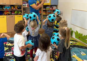 Zdjęcie przedstawiające prowadzącego zajęcia sportowe i małe dzieci trzymające niebieskie Piłeczki
