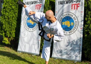 Instruktor taekwondo w białym stroju sportowym wykonuje wysokie kopnięcie na trawie. W tle stoją banery klubu taekwondo.