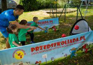 Dzieci w zielonych koszulkach uczestniczą w zabawie ruchowej z prowadzącym. Próbują trafić czerwonymi piłkami do małej bramki sportowej. W tle widoczny jest baner "Przedszkoliada.pl".