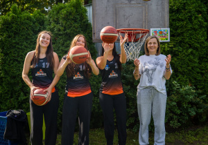 Cztery kobiety stoją przy koszu do koszykówki. Trzy z nich mają sportowe stroje i trzymają piłki do koszykówki, a jedna stoi obok nich w jasnym stroju.