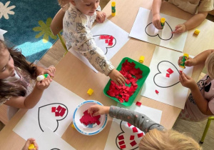 Dzieci pracują przy stoliczku, pokrywają serduszka kawałkami bibuły.
