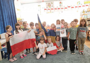 Część dzieci stoi, a część siedzi na dywanie w sali trzymając w rękach flagę i herb Aleksandrowa Łódzkiego oraz mapę Polski.