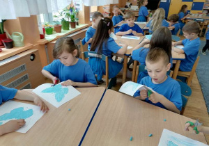 Dzieci siedzą przy stolikach i kolorują szablon motyla niebieską, pastelową kredką.
