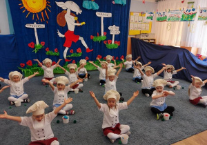 Dzieci z grupy II w przebraniu kucharzy siedzą na dywanie i podnoszą ręce do góry