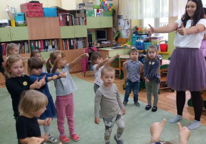 Dzieci tańczą z panią przy muzyce, mają wyciągnięte rączki przed sobą z kciukiem uniesionym do góry.