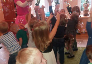 Grupa dzieci bawi się bańkami mydlanymi, próbują złapać bańki unoszące się w powietrzu.