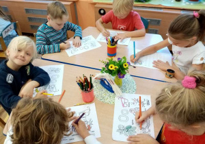 Sześcioro dzieci siedzi przy stoliku i wykonuje zadania (karty pracy) – kolorują obrazki, które w swojej nazwie maja na początku głoskę „o” oraz rysują szlaczki z małą i wielką literą „o” w liniaturze.
