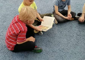 Chłopcy siedzą na dywanie i oglądają książkę „Z przygód Krasnala Hałabały”.