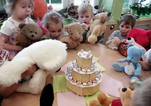 Grupka dzieci z misiami siedzi przy stoliku i zdmuchuje papierowe świeczki na papierowym torcie.