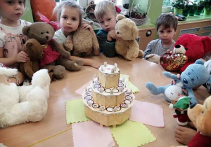 Grupka dzieci z misiami wokół papierowego tortu.