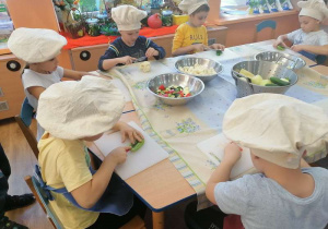 Dzieci siedzą przy stole i na deskach kroją warzywa na części.
