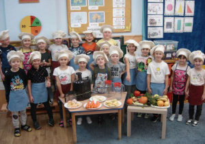 Grupa dzieci w fartuszkach i czapkach kucharskich a przed nimi warzywa i owoce.