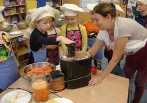 Dzieci wraz z nauczycielką wyciskają sok z marchewki przy użyciu sokowirówki.