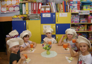 Pięcioro dzieci siedzi przy stoliku trzymając w dłoniach szklanki z sokiem z marchwi.