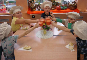 Pięcioro dzieci siedzi przy stoliku i stukają się szklankami z sokiem z marchwi.