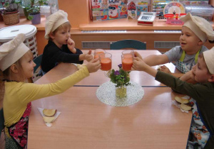 Czworo dzieci siedzi przy stoliku i stukają się szklankami z sokiem z marchwi.