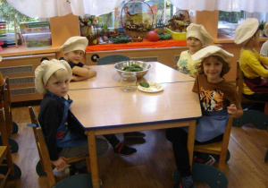 Czworo dzieci siedzi przy stoliku, na którym przygotowany jest słoik i produkty do kiszenia ogórków (ogórki, koper, chrzan, czosnek i liście z wiśni).