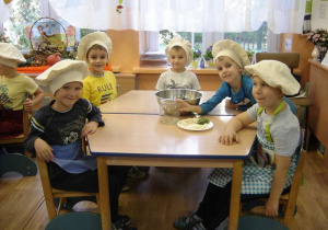 Pięcioro dzieci siedzi przy stoliku, na którym przygotowany jest słoik i produkty do kiszenia ogórków (ogórki, koper, chrzan, czosnek i liście z wiśni).