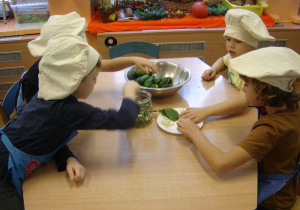 Czworo dzieci siedzi przy stoliku i wkłada do słoika produkty – składniki do kiszenia ogórków.