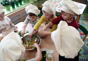 Grupka dzieci przy stolikach zapełnia słoje sałatką z warzyw.