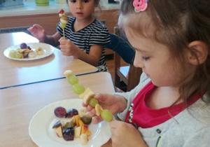 Dwie dziewczynki nabijają na patyczek do szaszłyków owoce – winogrona i jabłka