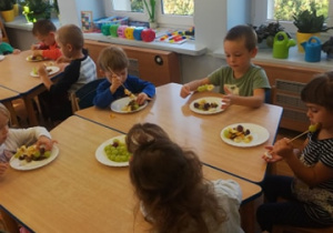 Grupa dzieci siedzi przy czwartym stoliku wykonuje owocowe szaszłyki – nakładając kawałki owoców na patyczki do szaszłyków