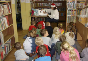 Grupa dzieci słucha opowiadania czytanego przez panią bibliotekarkę w czapce Mikołaja, która pokazuje ilustracje w książce.
