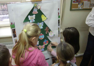 Trzy dziewczynki przyczepiają pokolorowane ozdoby na namalowaną zieloną choinkę.