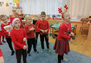 Chłopcy i dziewczynki stoją na dywanie śpiewając i ilustrując ruchem piosenkę pt. „Mikołaj z czerwonym noskiem”.