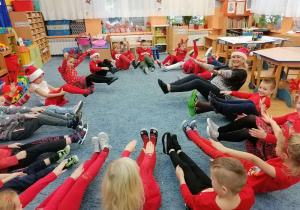 Podczas zajęć z języka angielskiego też było Mikołajkowo. Dzieci z panią Beatą siedzą na dywanie z uniesionymi nogami, ilustrując w ten sposób świąteczną piosenkę.