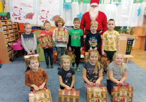 Dzieci stoją, siedzą na dywanie trzymając prezenty. Razem z nimi stoi Mikołaj.
