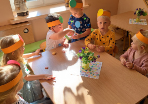 Dzieci w opaskach na głowach z kółeczkami siedzi przy stoliku i jedzą ciastka oraz gumy mamby.
