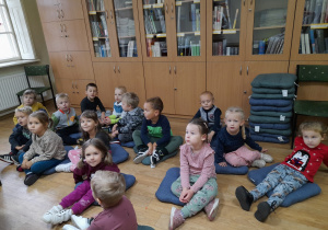 Dzieci siedzą na poduszkach w bibliotece słuchając opowiadania.