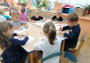 Dzieci siedzą przy stolikach i kolorują kredkami szablon znaku drogowego.