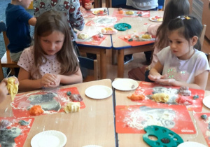 Dzieci siedzą przy stolikach i lepią owoce z masy solnej.